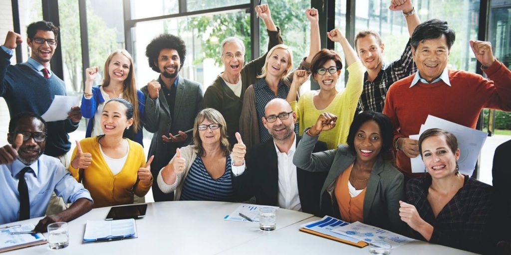 A multiethnic, multigenerational office team of mixed genders making thumbs-up and victory gestures for a group photo.