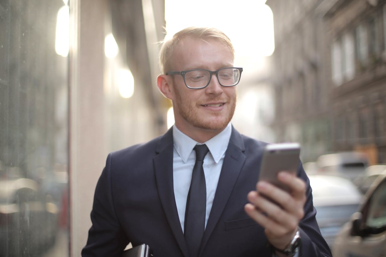 Businessman in a suit and tie smiling while looking at a smartphone, standing on a city street.