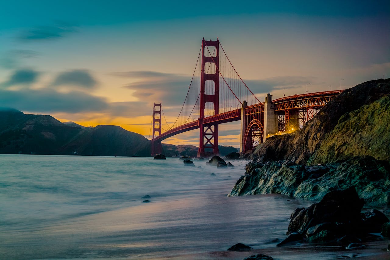 Golden Gate Bridge at sunset, viewed from a rocky shoreline with the ocean in the foreground.
